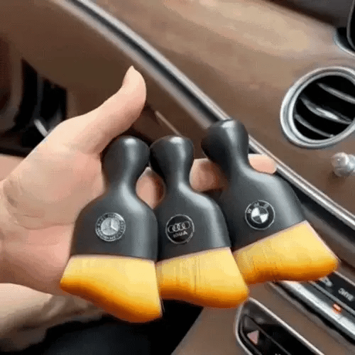 Hand holding three DustMate car detailing brushes with different car logos engraved on the handles, inside a car